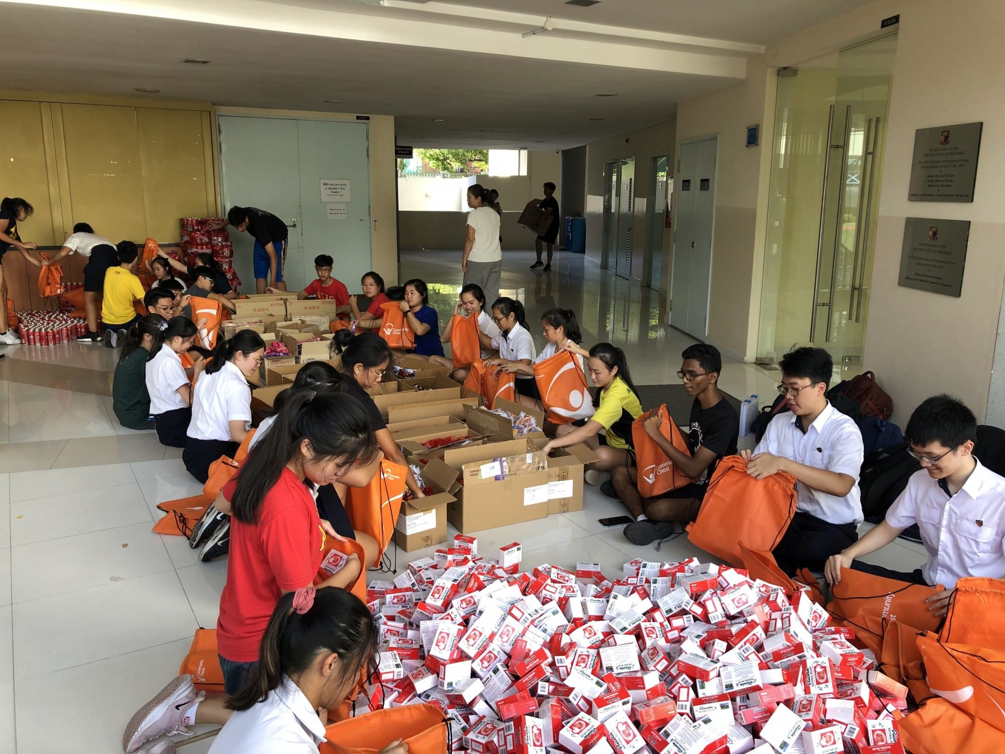 A large group of young volunteers sit on a floor packing red and white boxes into orange drawstring bags.